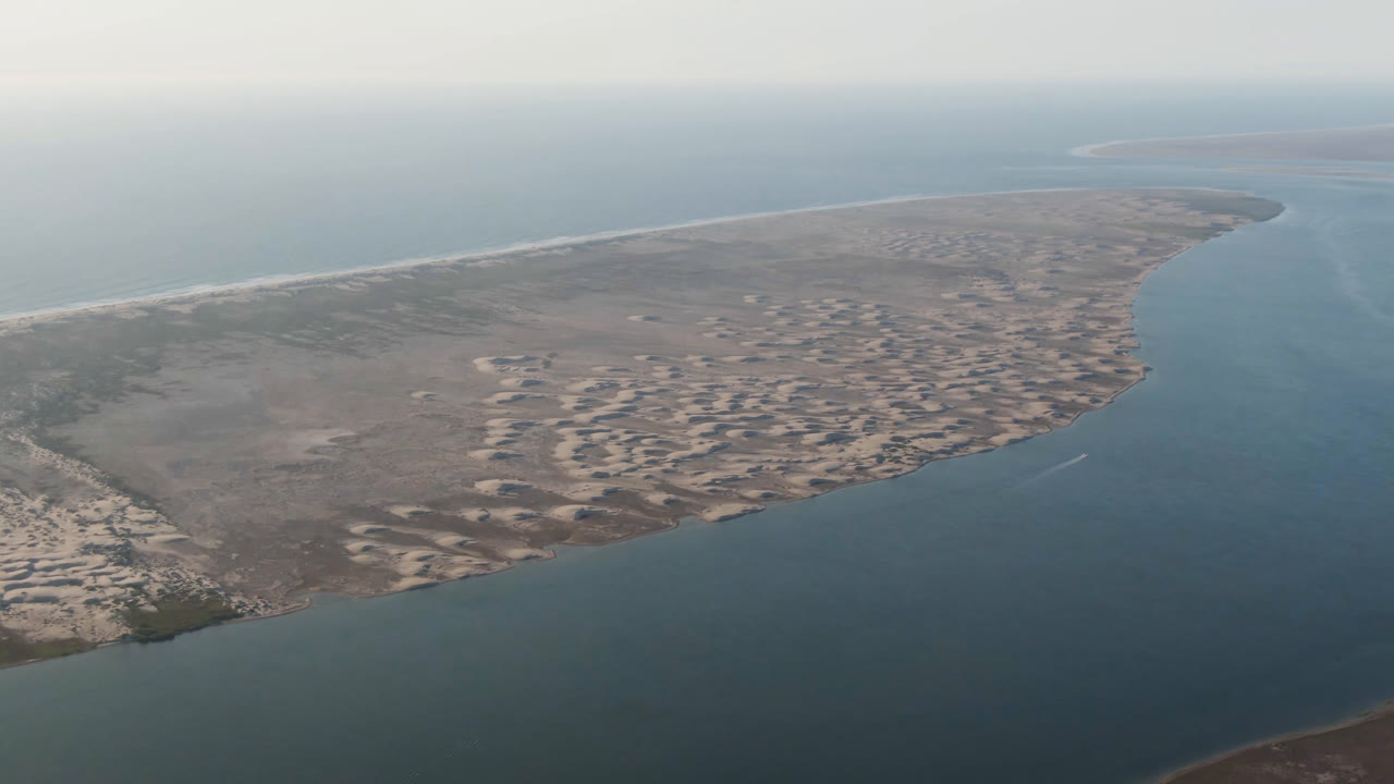 Opening aerial frame over Aldofo Lopez Mateos, Baja California Sur, with blue-green water in the foreground, pale sand banks along the shore, hazy sky, and no visible people.