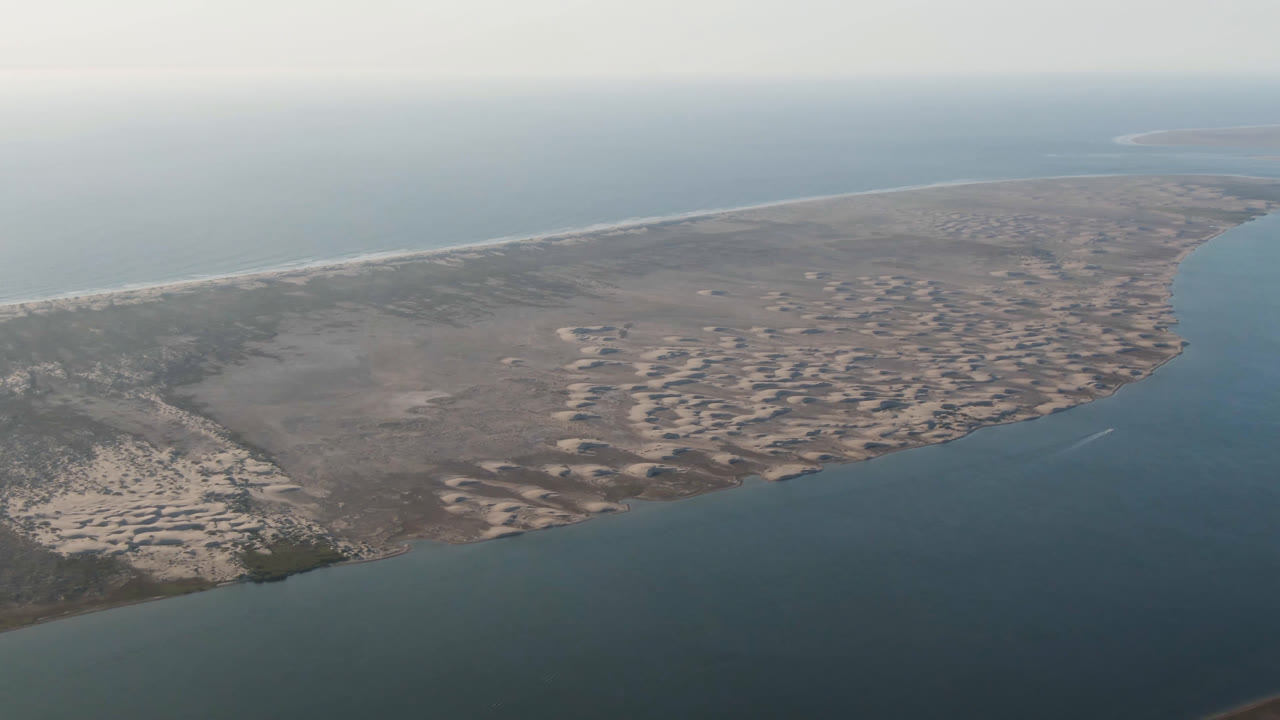 Opening aerial frame over Aldofo Lopez Mateos, Baja California Sur, with a sandy spit, dark scrubland, blue-green water, hazy horizon, and no visible people.