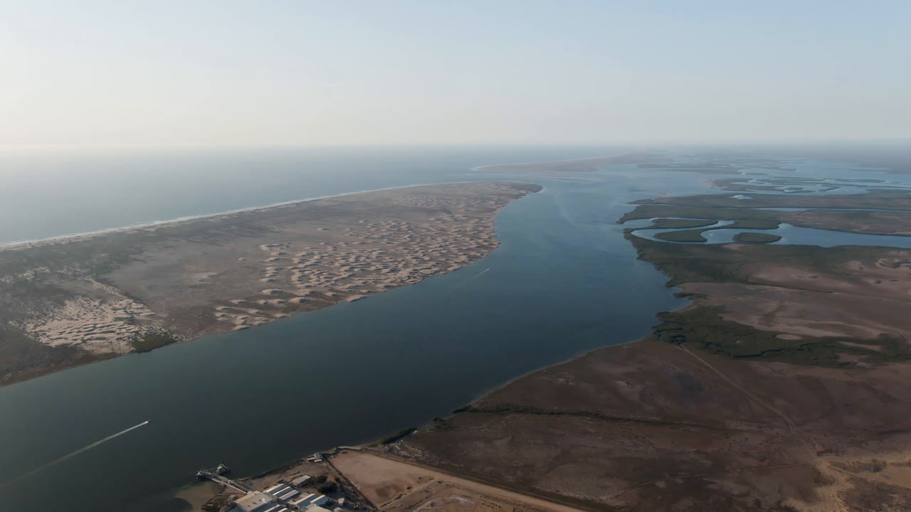 Opening aerial frame over Aldofo Lopez Mateos, Baja California Sur, with a broad water channel between a sandy ocean coast and darker wetland terrain, no visible people.