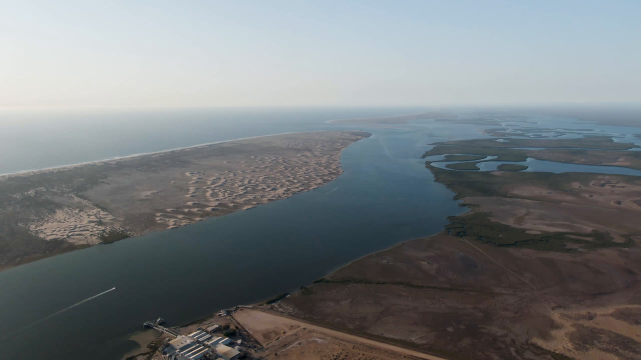 Aerial frame in Aldofo Lopez Mateos showing blue water running past a sandy coastal strip, ocean surf on the left, wetland pools on the right, and no visible people.