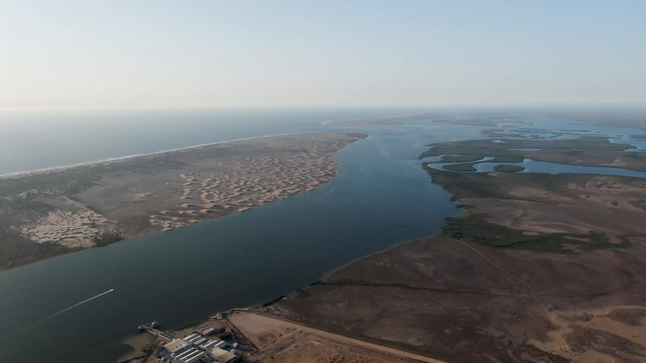 Wide drone view across a coastal channel with beach, town edge, and wetlands.