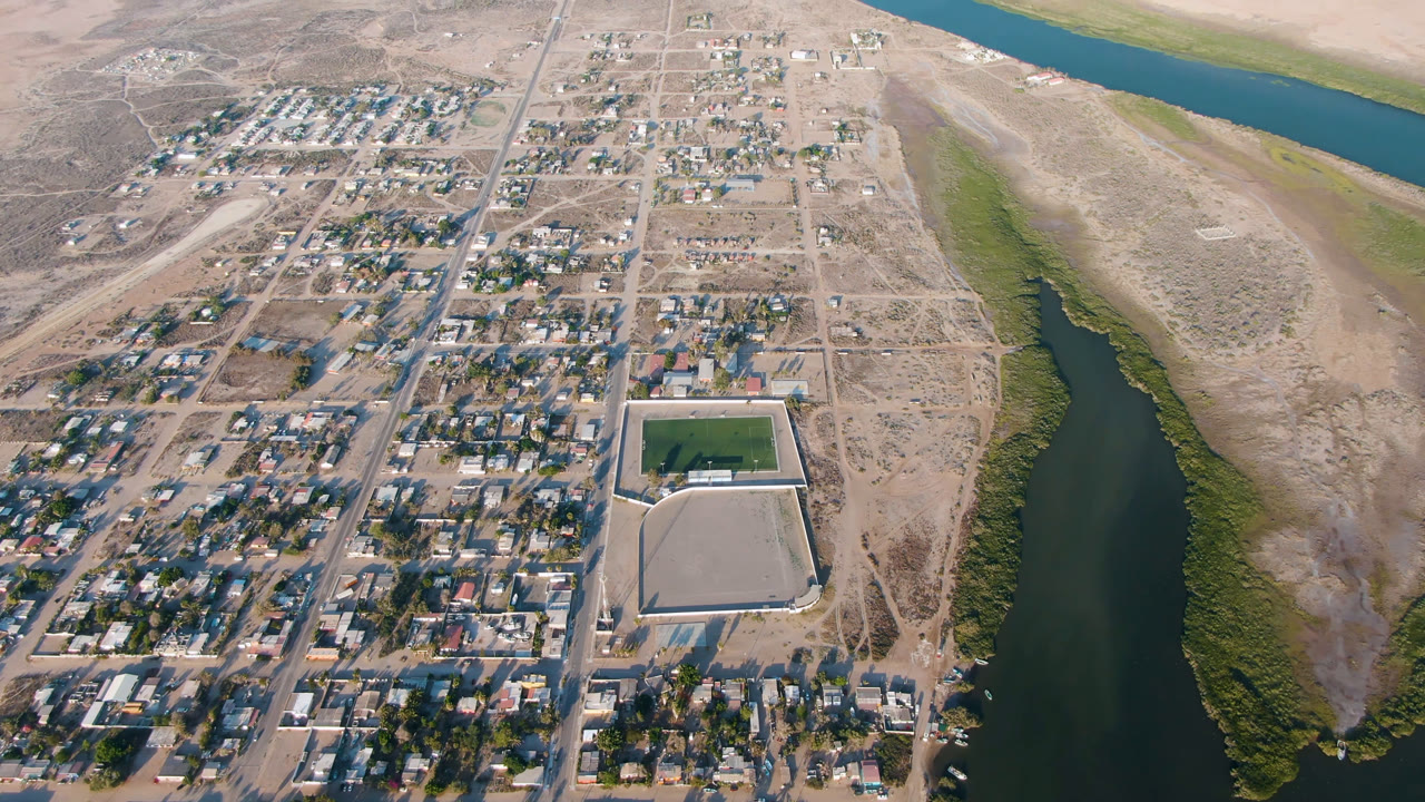 Second aerial frame of Adolfo Lopez Mateos, Mexico, showing the central road, scattered houses, a rectangular green sports field, river water to the right, and no visible people.