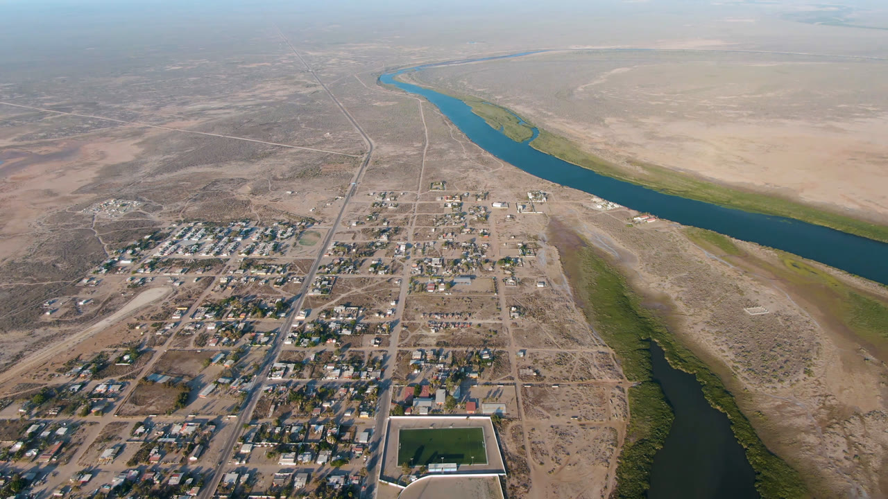 Aerial frame over Aldofo Lopez Mateos showing straight roads, clustered homes, the river curving through dry land, and no visible people.