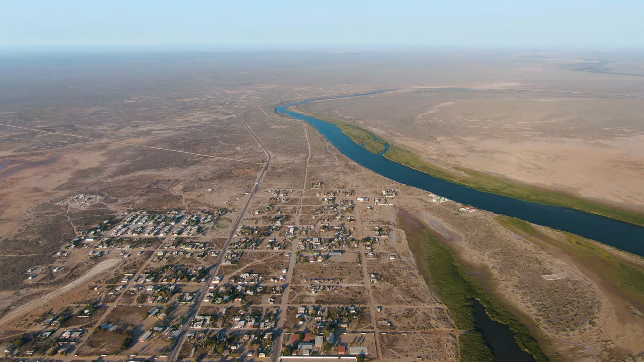 High aerial view over Aldofo Lopez Mateos, Baja California Sur, with a small town grid in the foreground, a curving river to the right, dry flats beyond, and no visible people.