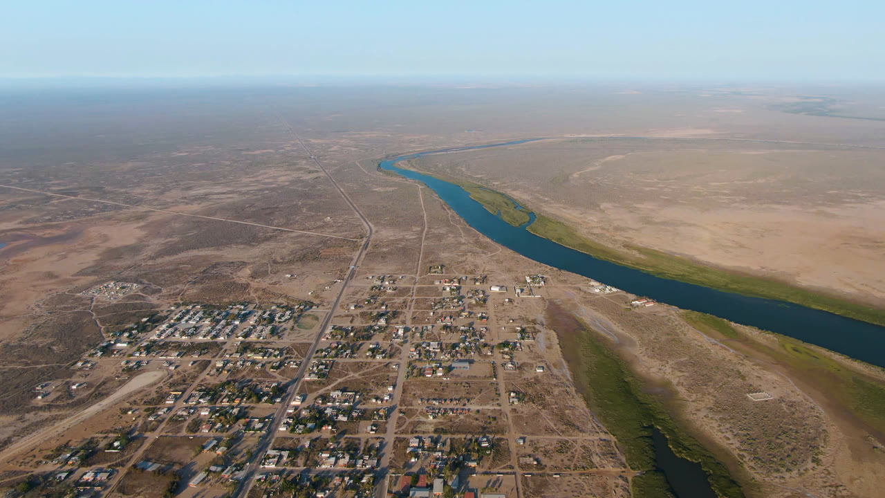 Aerial frame from Aldofo Lopez Mateos showing the town blocks slightly farther along the river bend, with tan open land on both sides and no visible people.