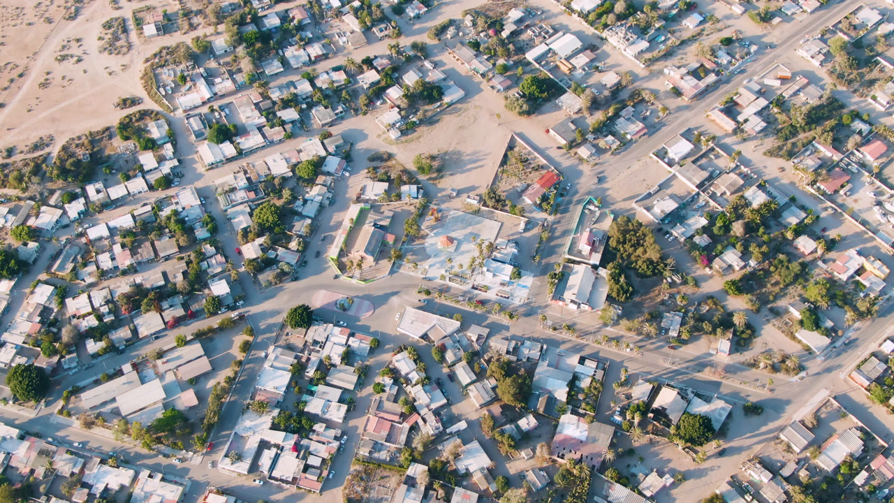 Overhead aerial frame of Aldofo Lopez Mateos, Baja California Sur, showing low-rise homes, pale sandy roads, trees, and walled lots with no visible people.
