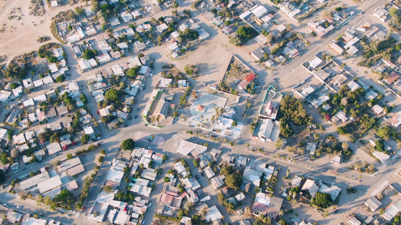 Aerial frame over Aldofo Lopez Mateos with crossing sandy roads, compact residential rooftops, scattered trees, and no visible people in the streets.