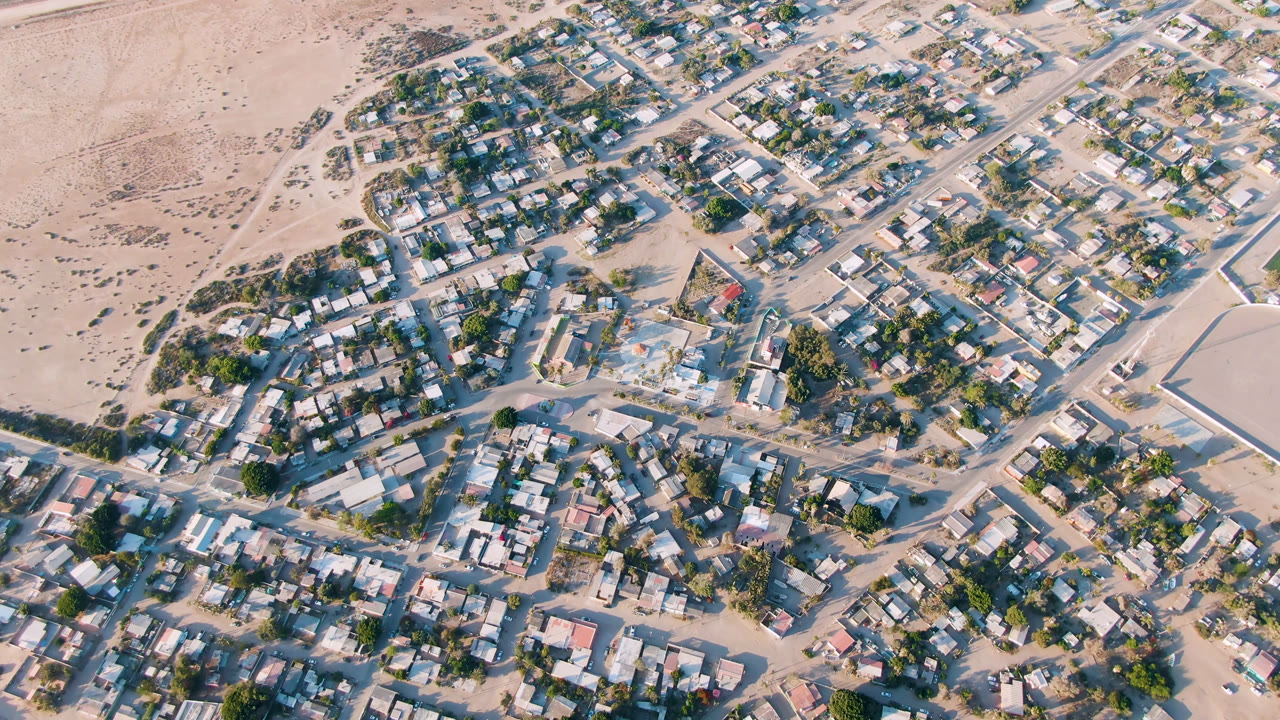 Second overhead frame of Aldofo Lopez Mateos, with the residential blocks slightly shifted across frame, showing more rooftops, tree cover, sandy streets, and no visible people.