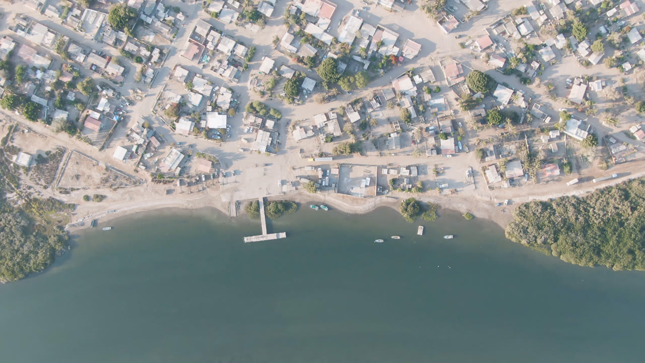 Aerial frame over Adolfo Lopez Mateos, Mexico, showing low buildings, pale dirt roads, a small dock, shoreline shrubs, river water, and no visible people.