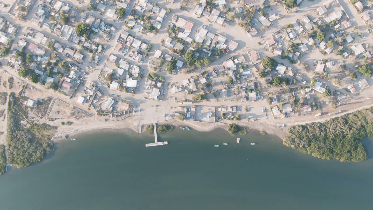 Aerial frame over Adolfo Lopez Mateos, Mexico, with the small pier near center, dense low buildings above the shore, green vegetation at the waterline, and no visible people.