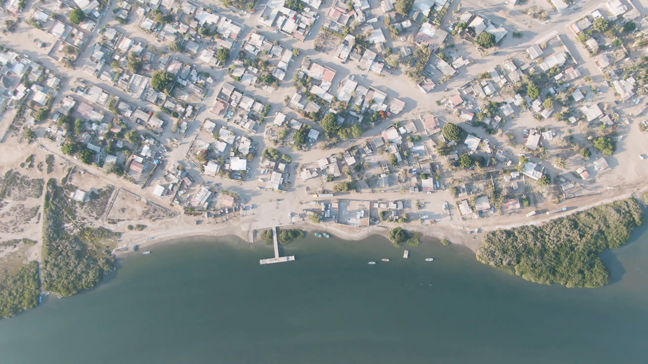 Aerial frame over Adolfo Lopez Mateos, Mexico, showing a compact settlement above the shoreline, a narrow dock extending into green water, small boats offshore, and no visible people.