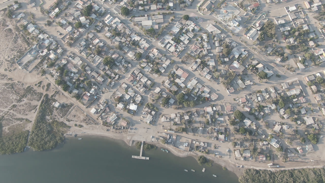 Drone view over Adolfo Lopez Mateos, Mexico, with beige rooftops, dirt streets, a shoreline road, a narrow pier near the lower center, and no visible people.