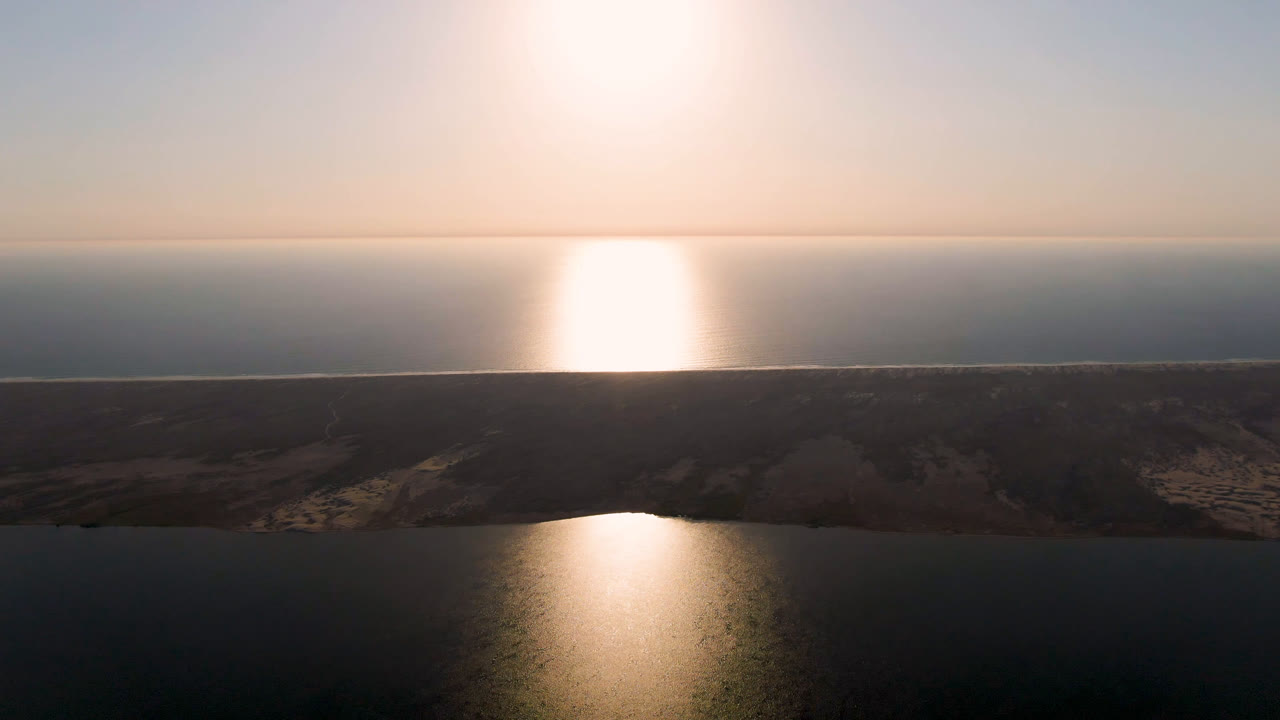 Aerial frame over Aldofo Lopez Mateos, Baja California Sur, with bright reflected light on water, a dark sandbar across the middle, pale sky above, and no visible people.