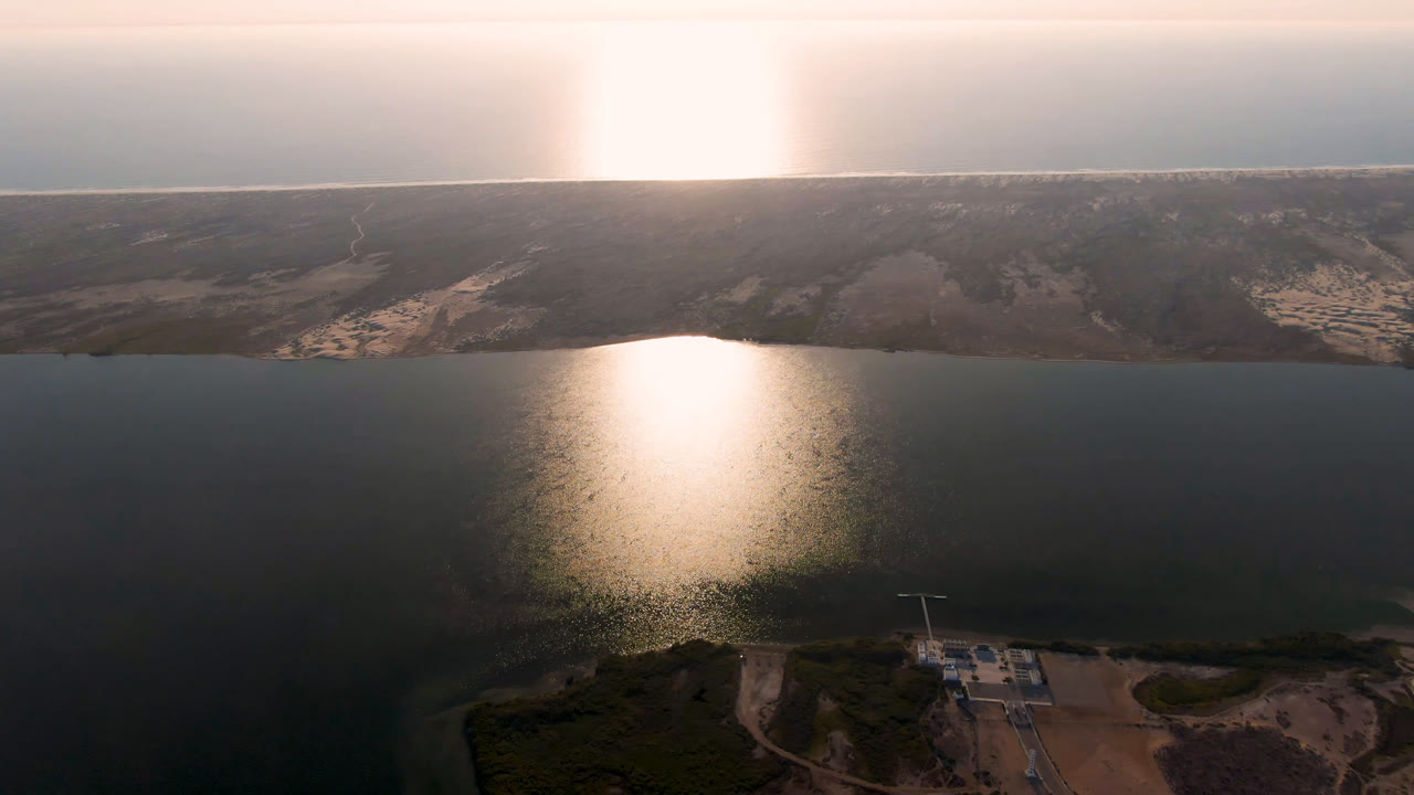 Aerial view over dark water with sun glare and a sandy shoreline on the horizon.