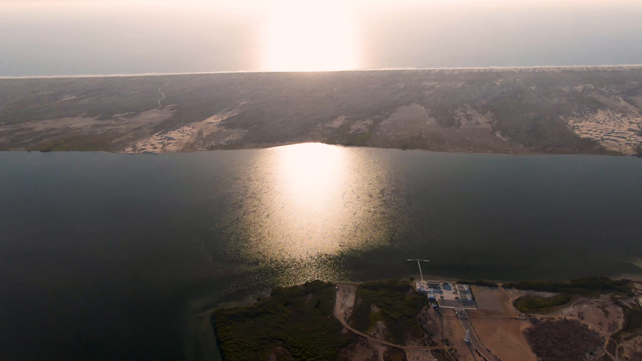 Aerial frame over Aldofo Lopez Mateos water, with the sun glare centered on the surface, scrub and small shoreline structures below, and no visible people.