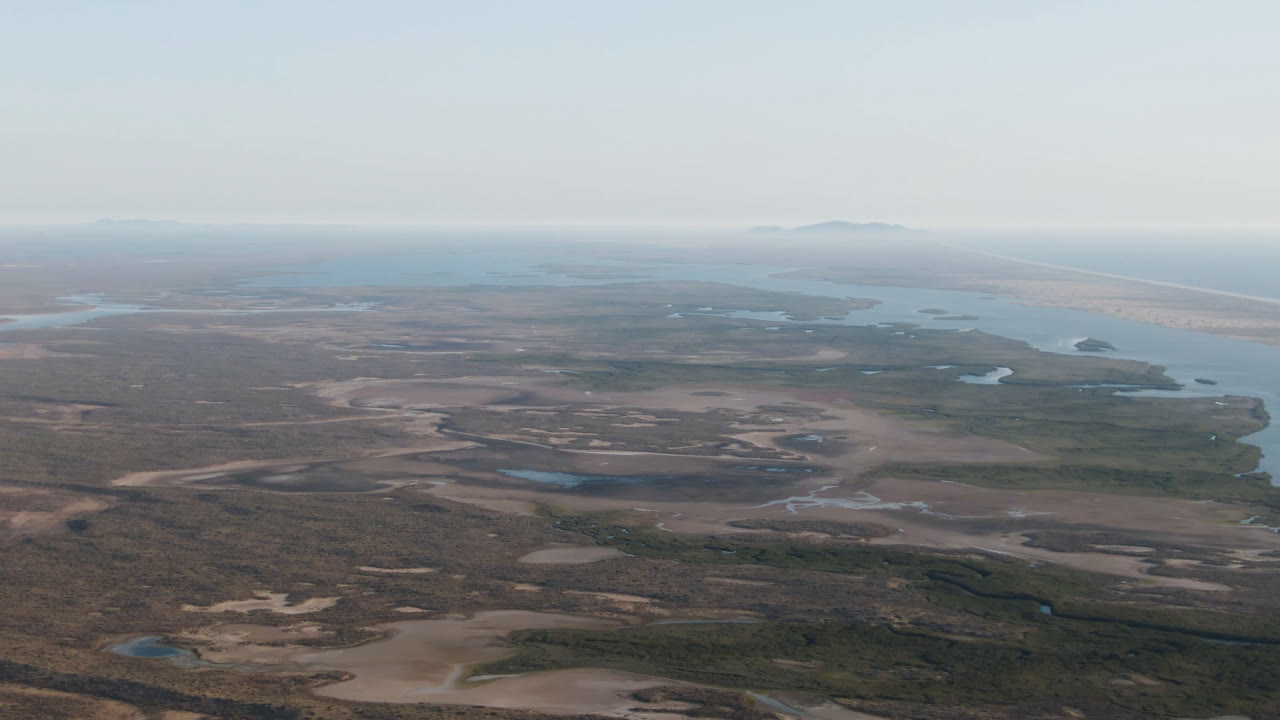 Wide aerial view over Aldofo Lopez Mateos, Baja California Sur, with sandy flats in the foreground, lagoon water and ocean beyond, distant land on the horizon, and no visible people.