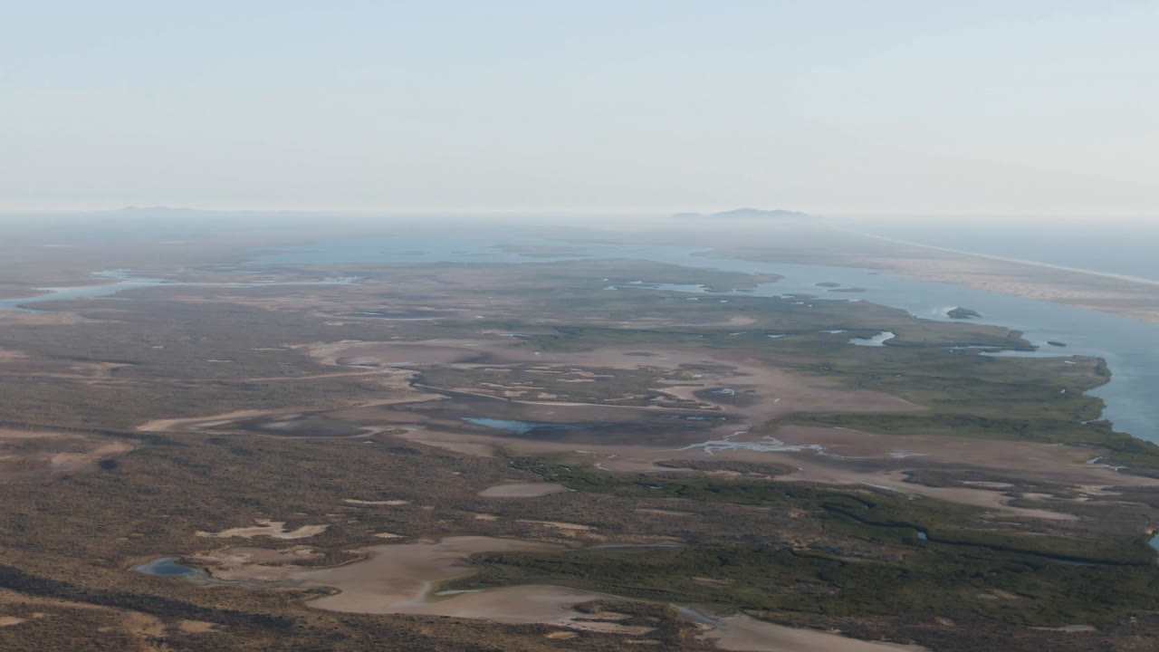 Aerial frame over Aldofo Lopez Mateos, Baja California Sur, showing pale dune patches, dark scrub, small water pockets, a long beach line, ocean, and no visible people.
