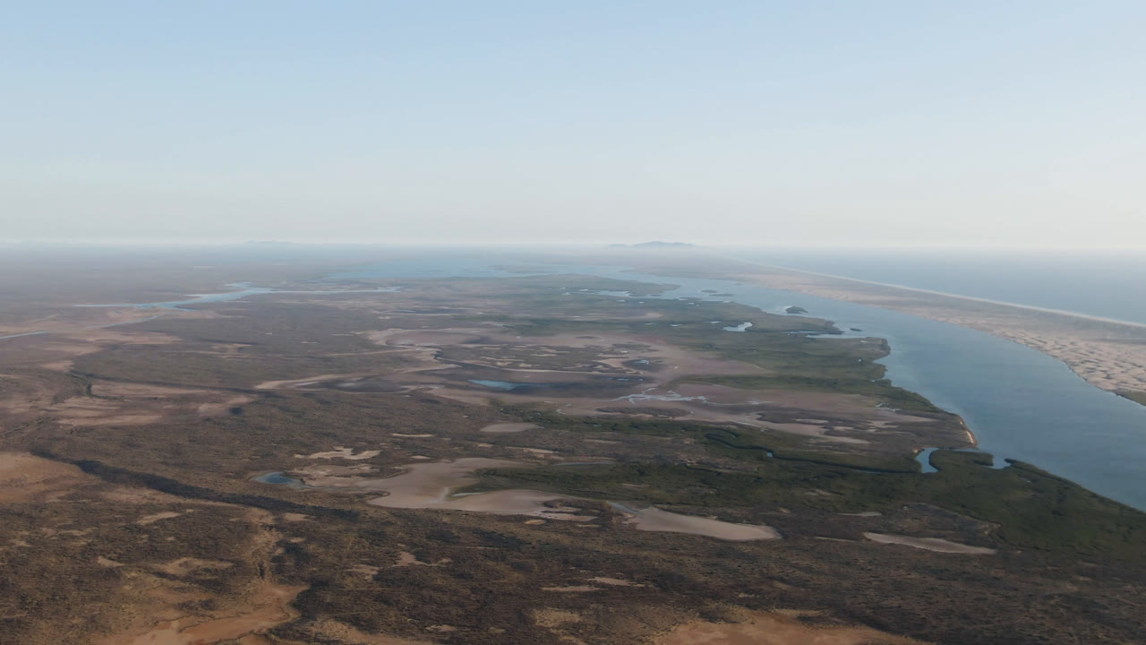 Wide aerial frame over Aldofo Lopez Mateos, Baja California Sur, with dry coastal flats in the foreground, a blue water channel at right, pale beach and ocean beyond, and no visible people.
