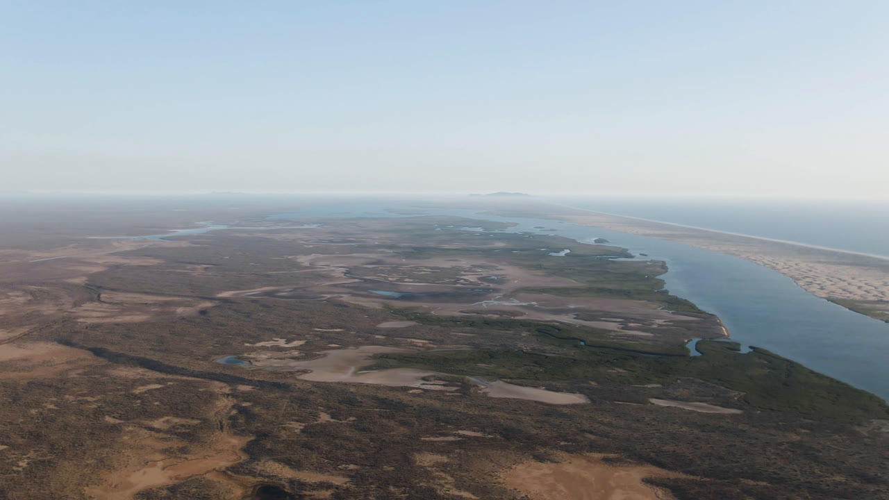 A second aerial view over Aldofo Lopez Mateos shows more of the brown and green flats below, the water channel curving along the right side, the long beach strip, and no visible people.