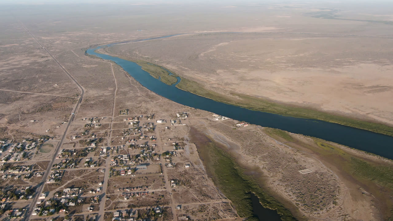 Aerial frame over Adolfo Lopez Mateos, Mexico, with the river running diagonally across the scene, low buildings clustered below it, dry roads, open flats, and no visible people.