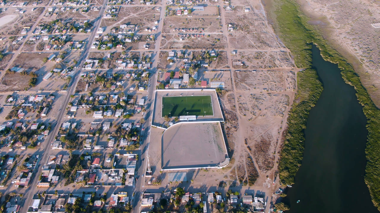 Opening aerial frame over Aldofo Lopez Mateos, Baja California Sur, with gridded homes, a green sports field, pale walled grounds, scrubland, and a dark river channel at right, with no visible people.