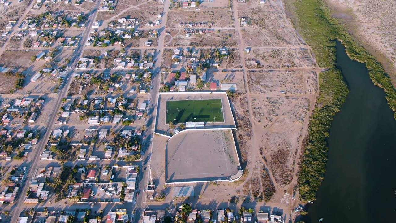 Second aerial frame in Aldofo Lopez Mateos, Baja California Sur, showing more of the residential grid on the left, the green field and pale walled area near center, and the river margin on the right, with no visible people.