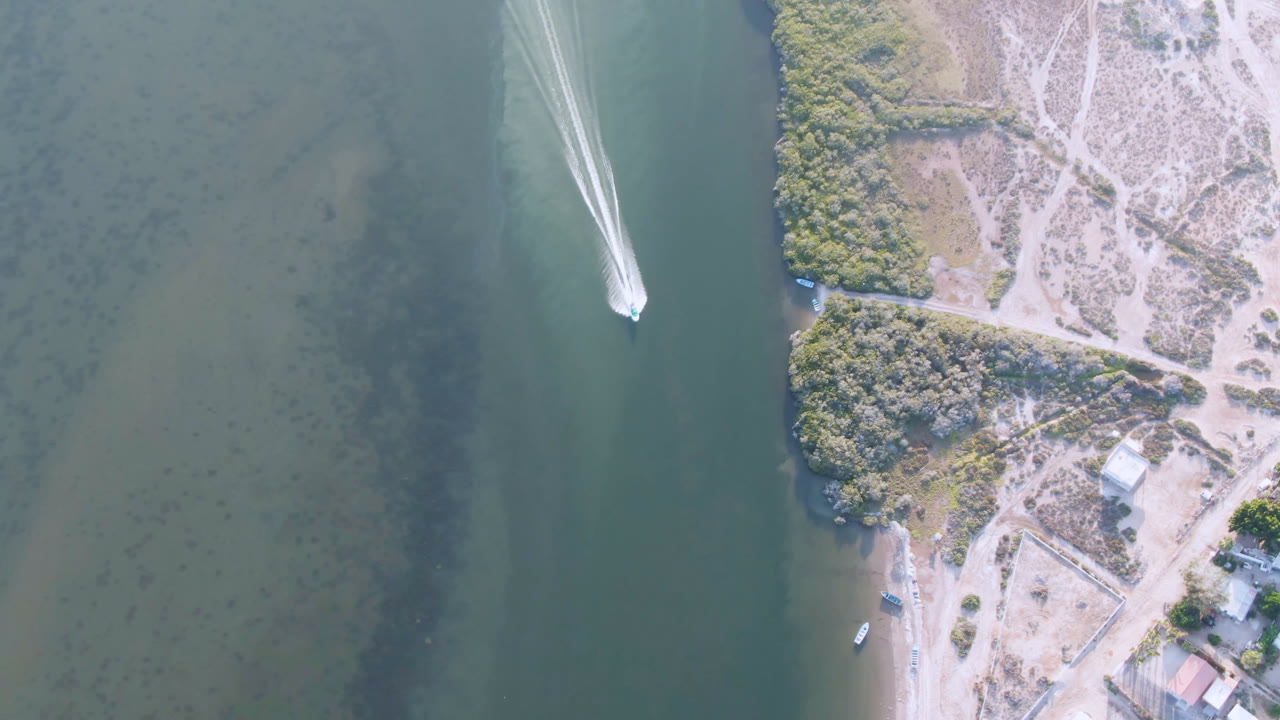 Overhead view of a small boat on green water beside a sandy shoreline.