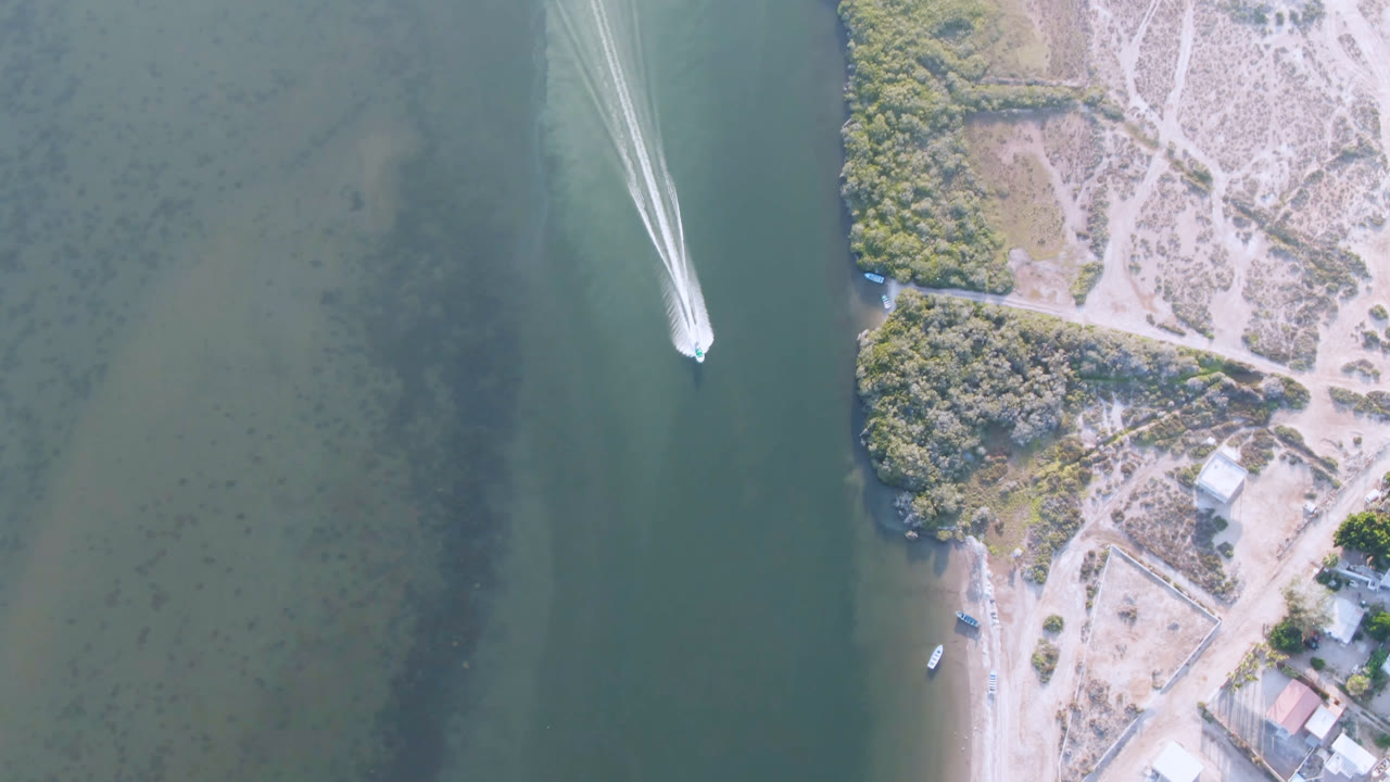 At 0:01, the boat has advanced across the green channel in Aldofo Lopez Mateos, with a narrow white wake, pale sand, sparse vegetation, and shoreline structures visible to the right, no visible people.