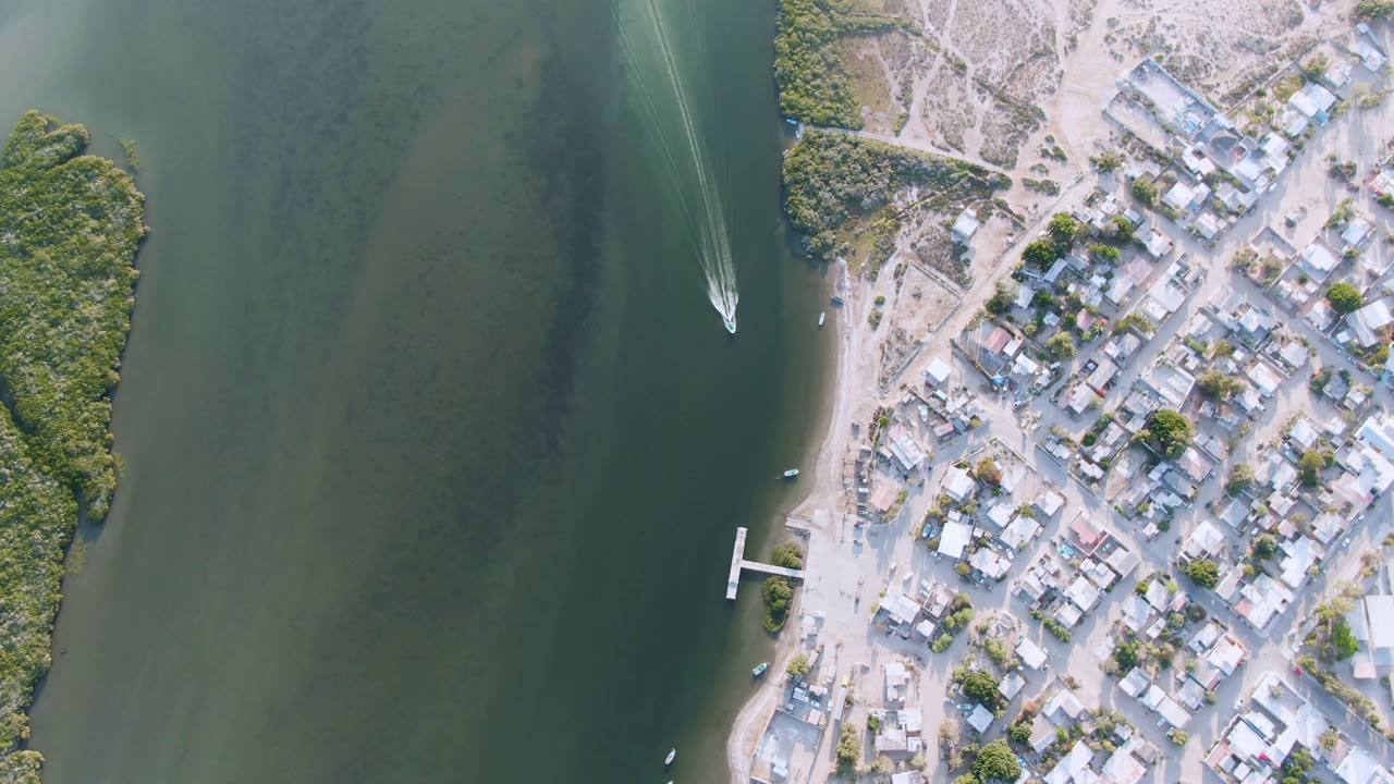 Overhead aerial frame of Aldofo Lopez Mateos, Baja California Sur, with green water on the left, clustered buildings on the right, a dock near the shore, a small boat wake near the upper water, and no visible people.