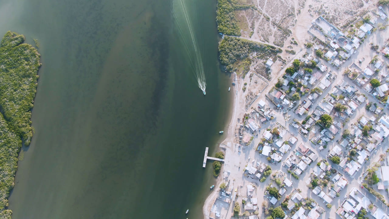 Aerial view over Aldofo Lopez Mateos, Baja California Sur, showing the boat wake slightly farther down the green water, pale roads crossing the town, a shoreline dock, trees, rooftops, and no visible people.
