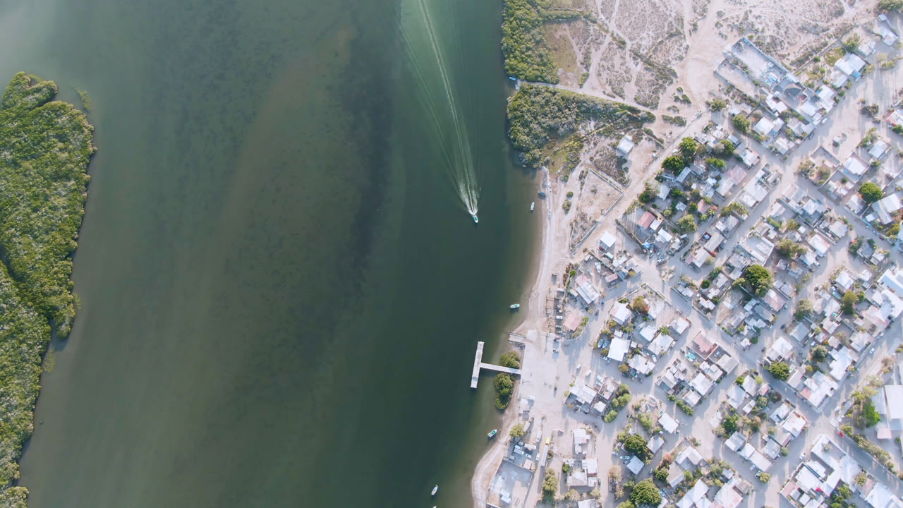 Top-down drone view of a town edge, dock, and boat wake on calm water.