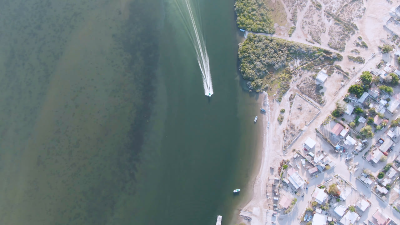 Overhead view of a white boat on green water beside a sandy town shoreline.