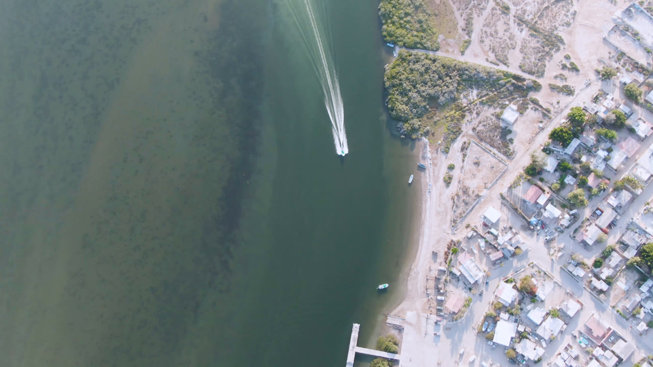 Aldofo Lopez Mateos aerial frame with the white boat slightly farther along the water channel, sandy bank and compact town blocks visible to the right, and no visible people.