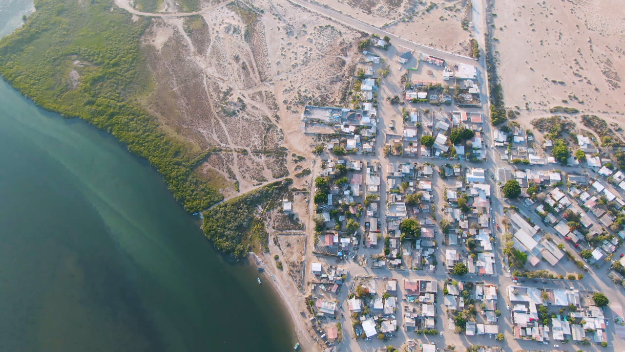 Aerial frame over Aldofo Lopez Mateos showing the water channel along the left, dense shoreline vegetation, low buildings, sandy streets, parked vehicles, and no visible people.