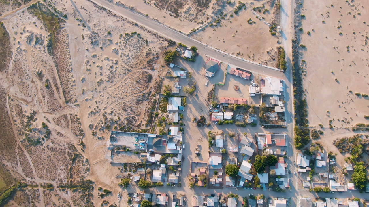 Aerial frame in Aldofo Lopez Mateos with compact homes, a diagonal road, dry open ground, scattered shrubs, and no visible people.