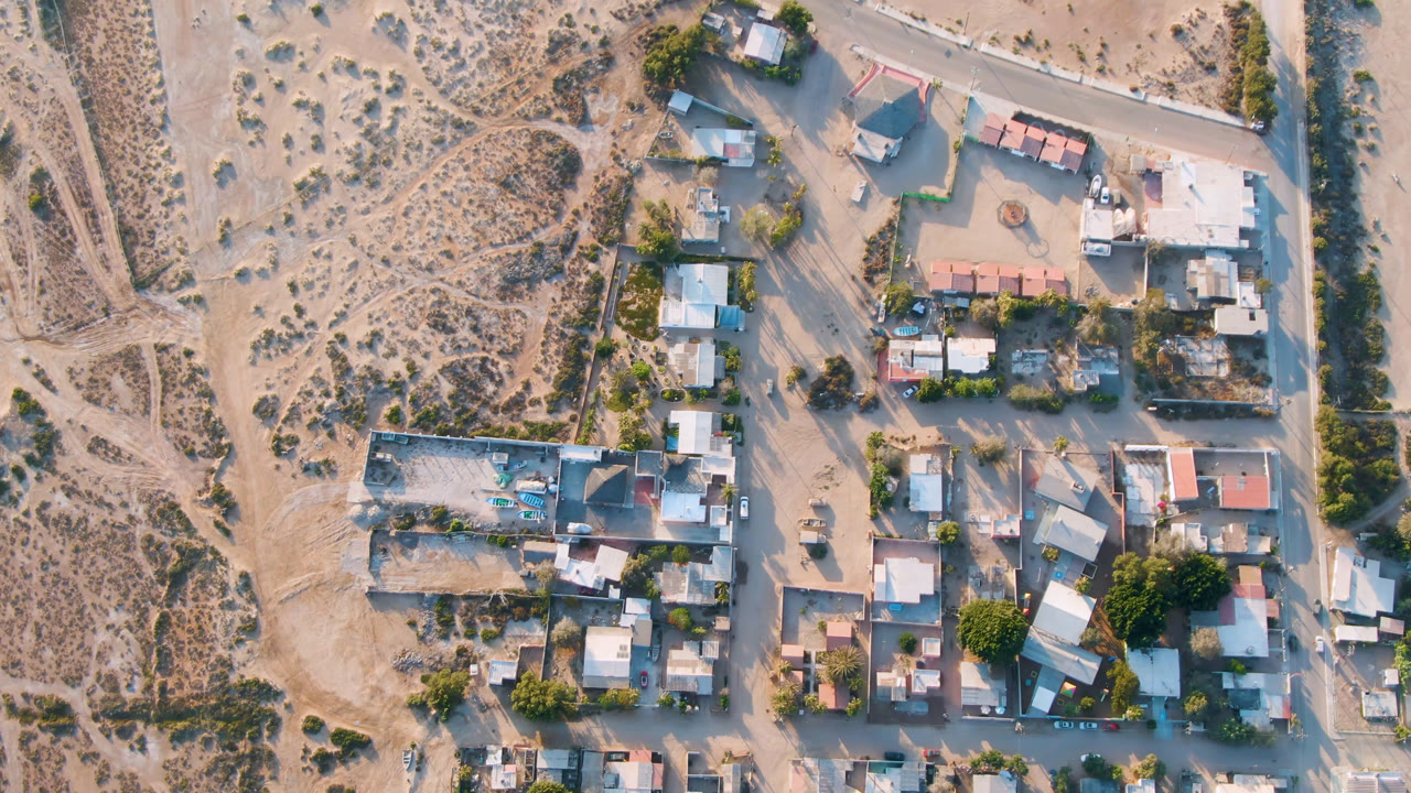 Second overhead frame in Aldofo Lopez Mateos with the residential cluster centered, a pale road along the upper edge, scrubland to the left, and no visible people.