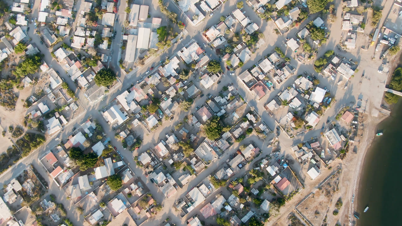 High aerial frame over Aldofo Lopez Mateos showing dense low buildings, intersecting pale roads, sparse greenery, the river edge at right, and no visible people.