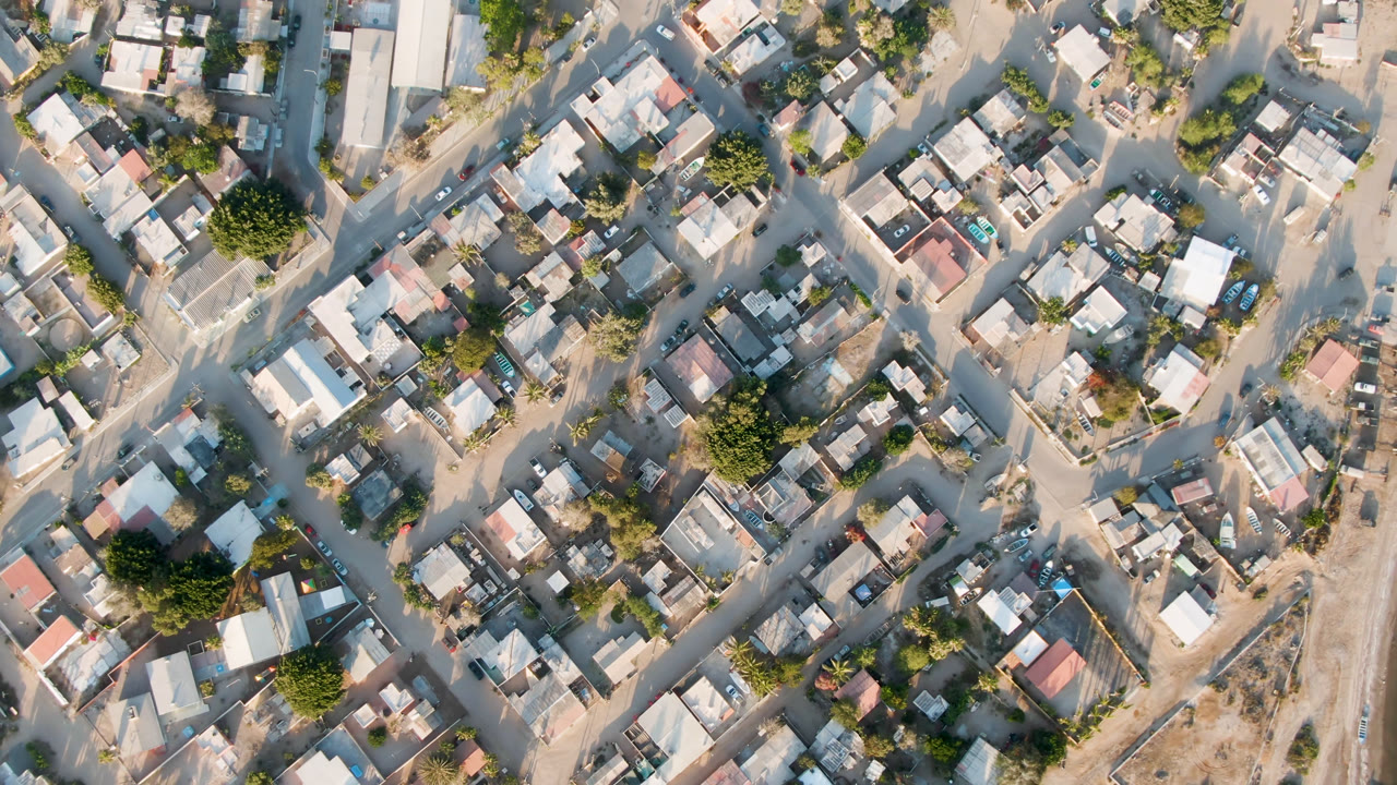 Overhead view in Aldofo Lopez Mateos with dense rooftops, crossing dirt roads, green trees, dry open lots, and no visible people.