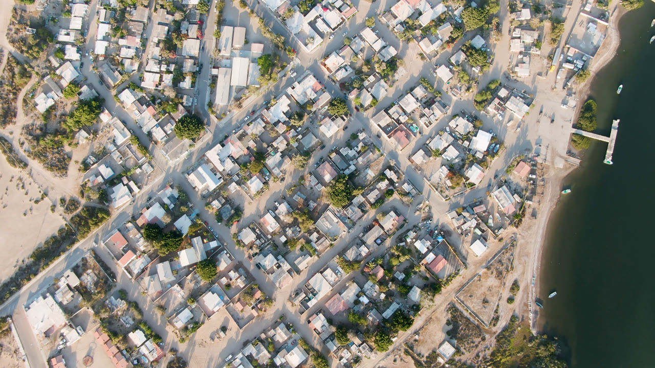 High aerial frame over Aldofo Lopez Mateos, Mexico, showing low buildings, sandy roads, trees, a beach edge, dark river water, a small dock, and no visible people.