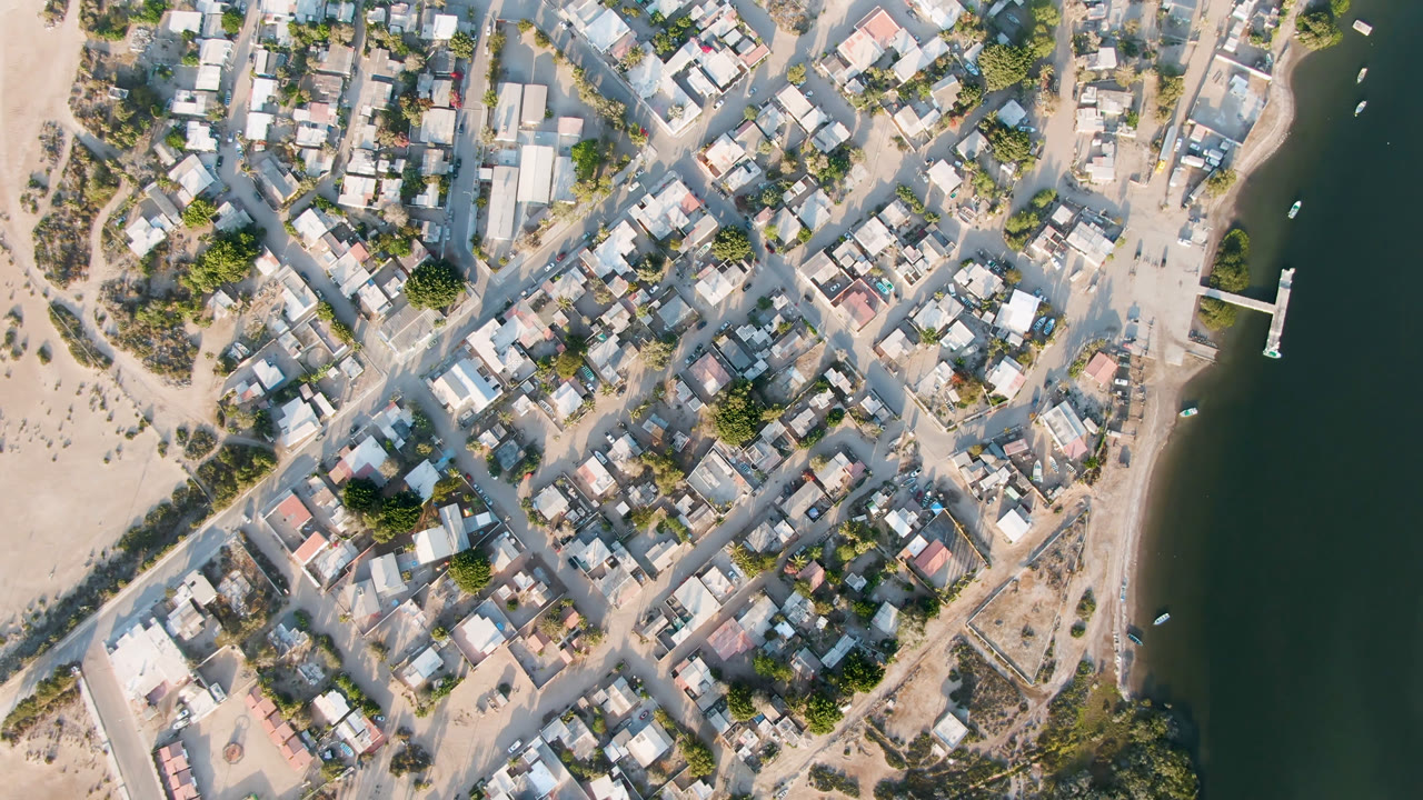 Top-down view of Aldofo Lopez Mateos with clustered rooftops, pale roads, scrub vegetation, a narrow shoreline, dark green river water, small boats, and no visible people.