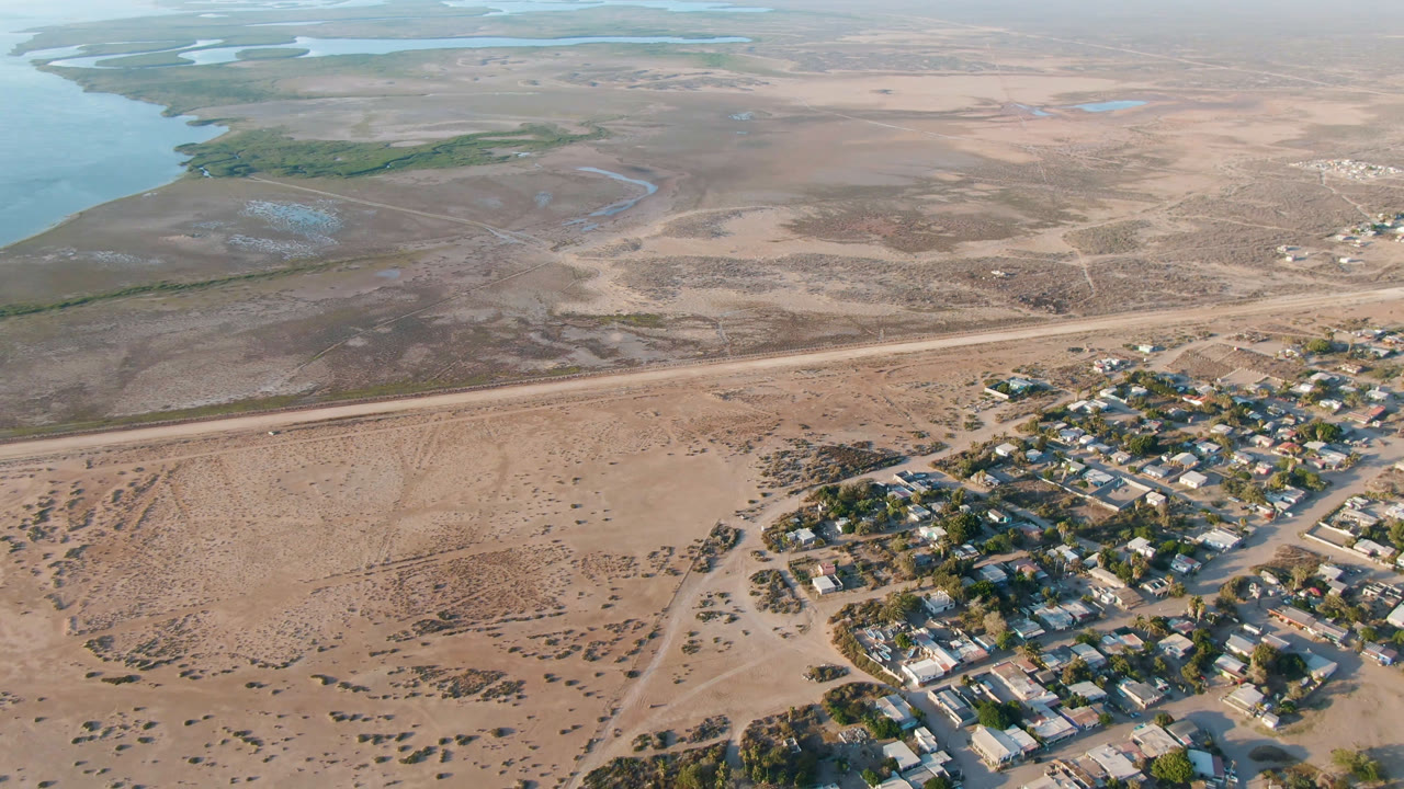 Aerial view in Adolfo Lopez Mateos, Mexico, showing the village shifted slightly farther right, a straight road across the sandy midground, river water on the left, and no visible people.
