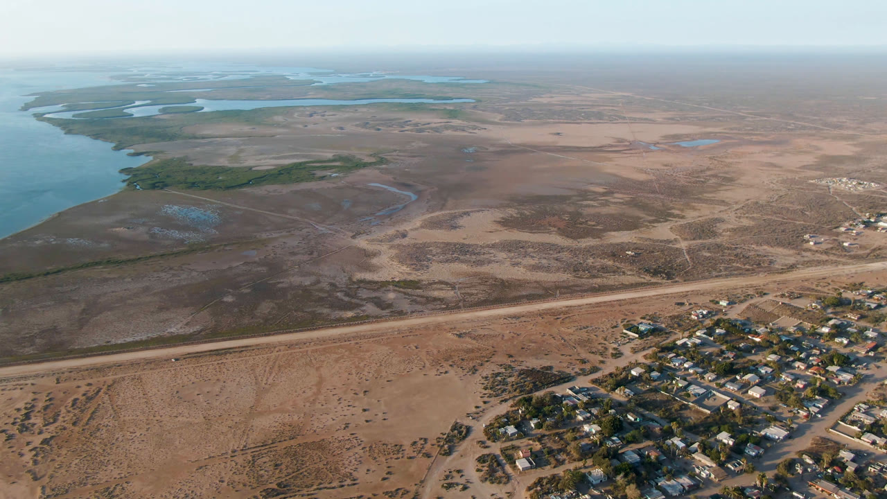 Wide aerial frame over Adolfo Lopez Mateos, Mexico, showing blue coastal water at left, tan undeveloped land, dirt roads, and a small settlement at lower right with no visible people.