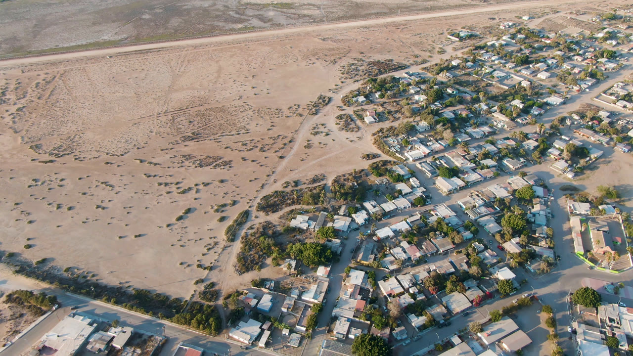 Second aerial frame of Aldofo Lopez Mateos with residential blocks filling the lower half, scrubby sandy terrain in the middle, and no visible people on the roads.