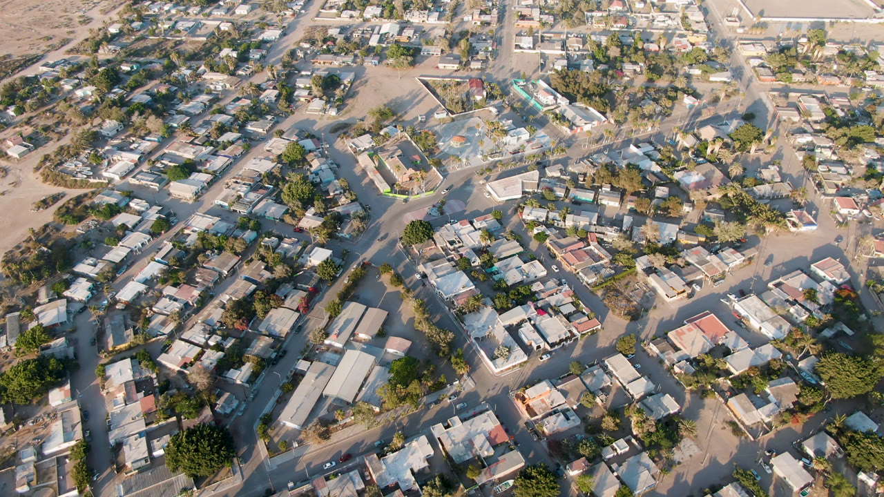 A high aerial view over Aldofo Lopez Mateos shows a curving road through low buildings, trees, parked vehicles, and dry open land, with no visible people.