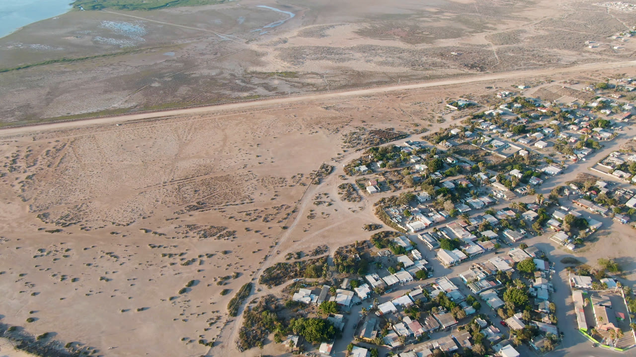 One second into the clip, the aerial view keeps Aldofo Lopez Mateos housing at the lower right while sandy scrubland and faint vehicle tracks fill the middle ground, with no visible people.