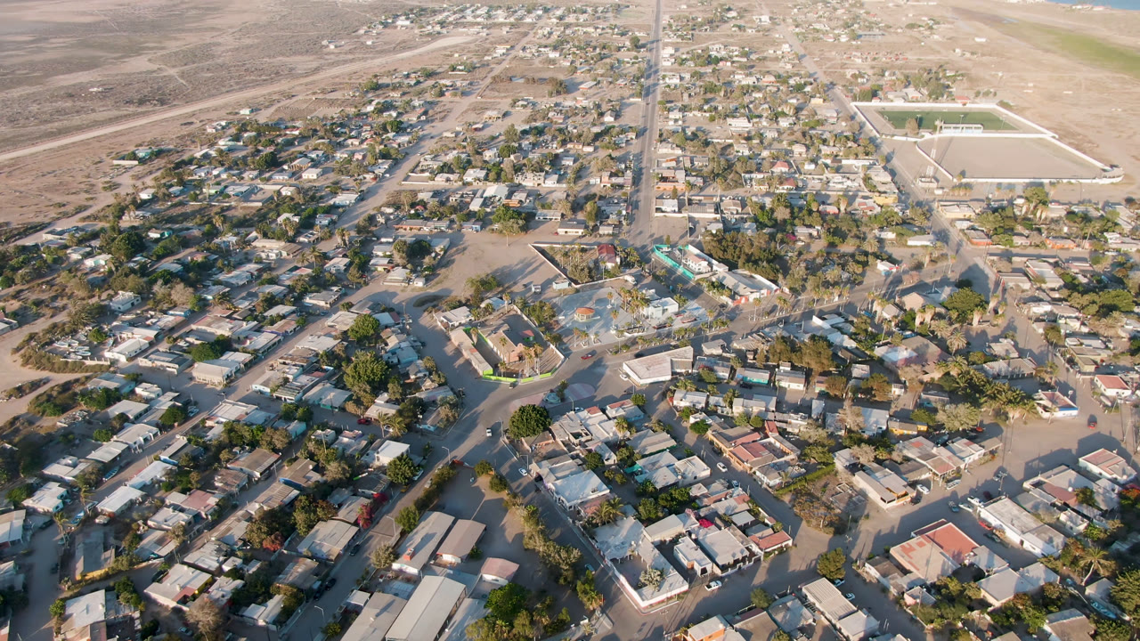 Second aerial frame over Aldofo Lopez Mateos with the central neighborhood slightly more prominent, showing a rectangular sports field to the right, desert beyond town, distant water, and no visible people.