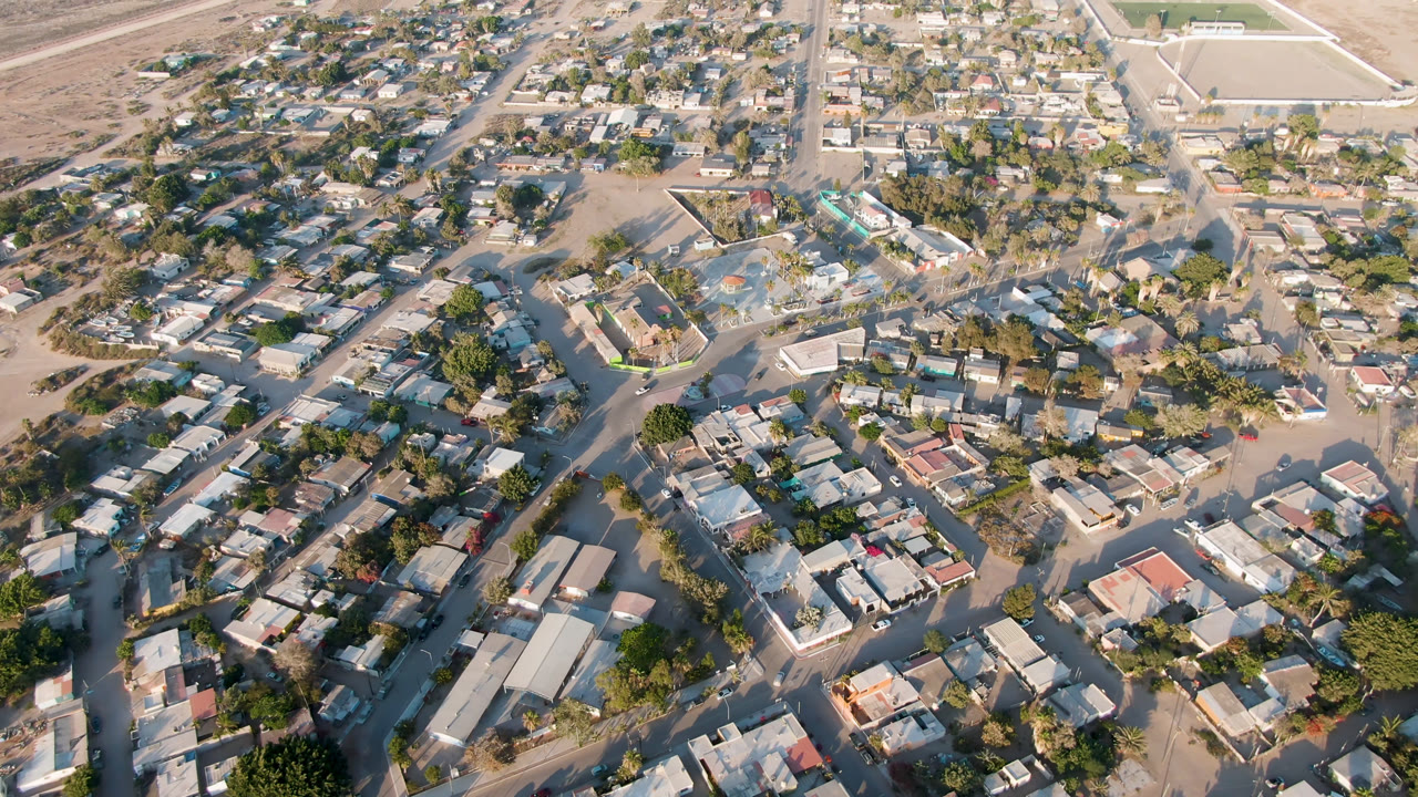 Aerial frame in Aldofo Lopez Mateos with compact homes, green trees, intersecting dusty streets, pale waterlogged patches, and no visible people.