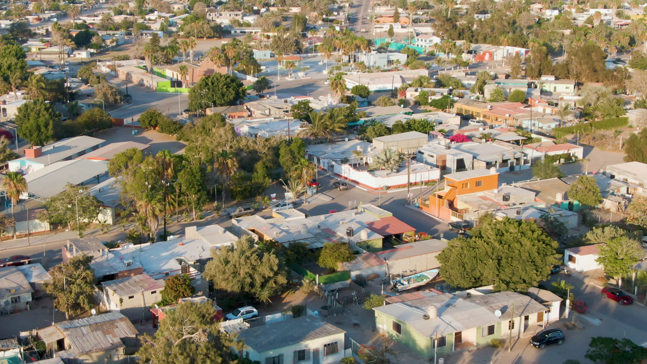 Opening aerial frame over Comondu, Mexico, showing flat-roofed homes, palms, narrow streets, parked cars, and no visible people.