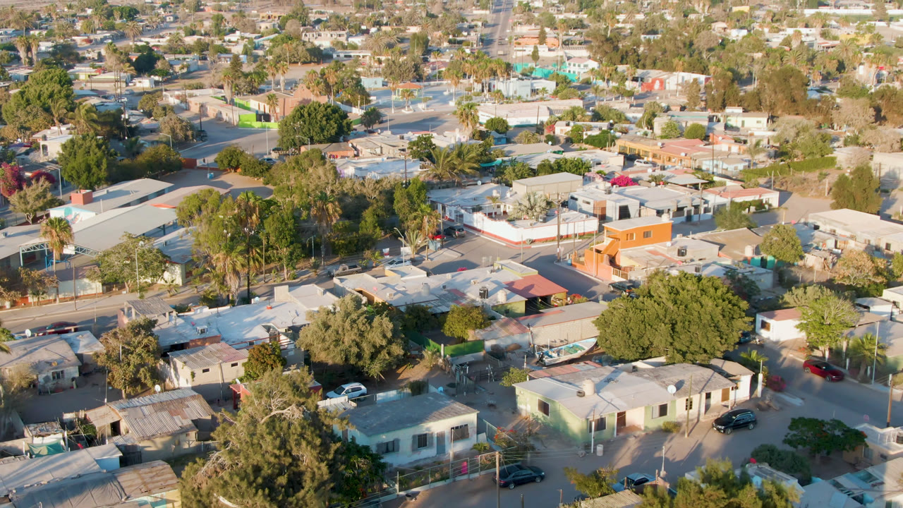 Second aerial frame over Comondu, Mexico, with beige rooftops, green trees, a few parked vehicles, and no visible people in the streets.