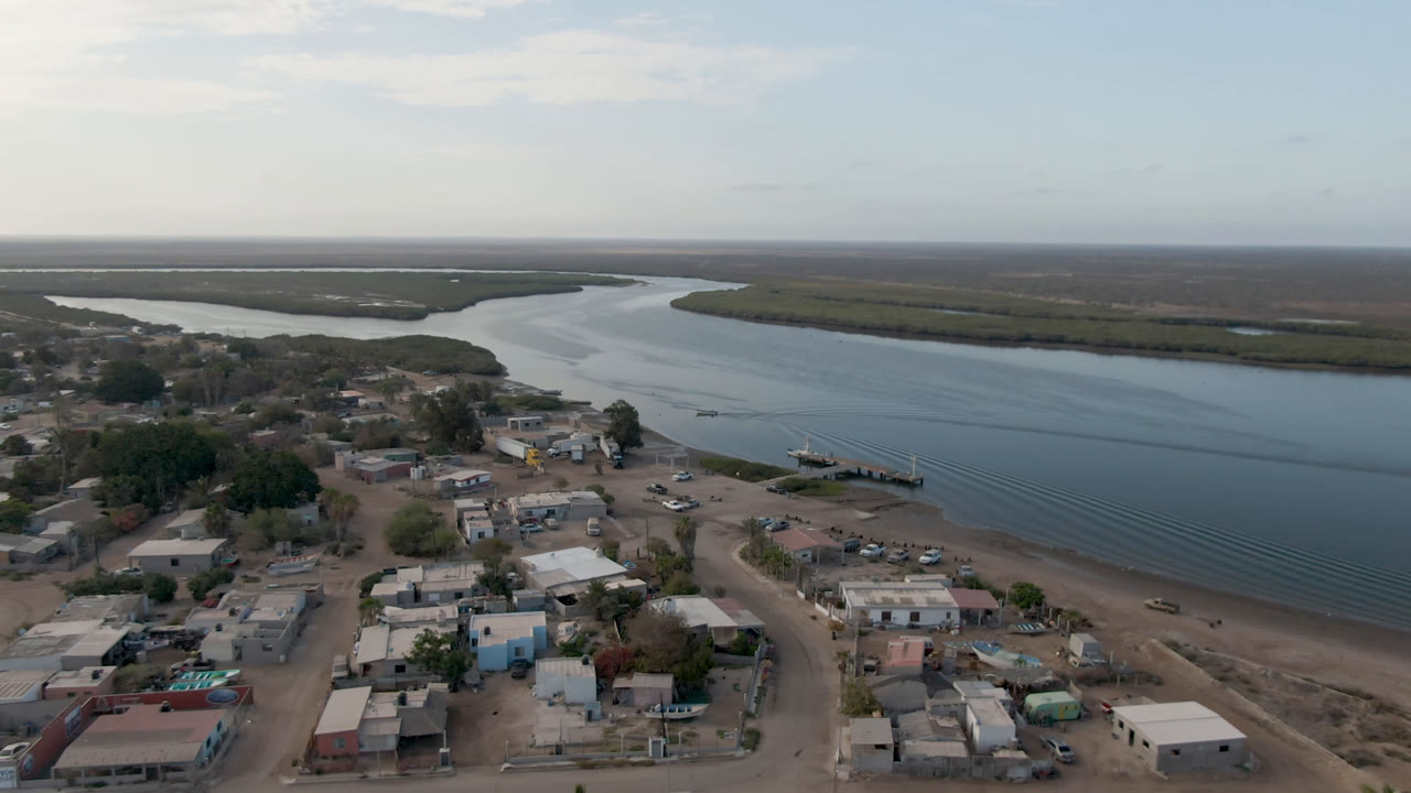 Aerial frame over Adolfo Lopez Mateos, Mexico, showing more of the shoreline dock, low buildings, dirt roads, broad blue-gray water, and no visible people.